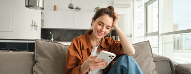 Portrait of happy young brunette woman sitting on sofa with smartphone, reading notification,...
