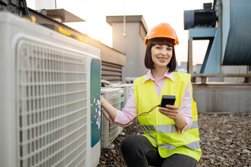 Beautiful repairwoman kneeling in front of air conditioner while holding digital multimeter in hands. Caucasian brunette with orange hard hat checking readiness of cooling system for installation.