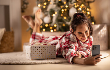 Cheerful woman in pajamas lying on carpet in living room with gift next to Christmas tree taking...