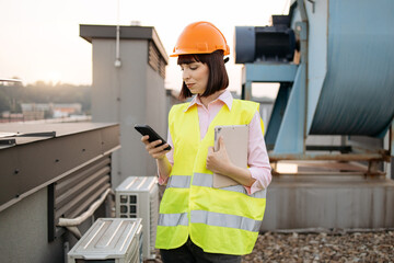 Front view of attentive caucasian civil engineer operating smartphone and tablet while standing on background of metal industrial pipes. Lady inspecting equipment with modern devices outdoors.