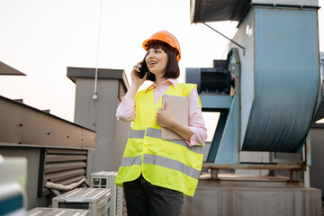 Factory female worker standing on rooftop with background of blue air duct and several metal boxes. Competent caucasian woman using modern smartphone and digital tablet for work outdoors.