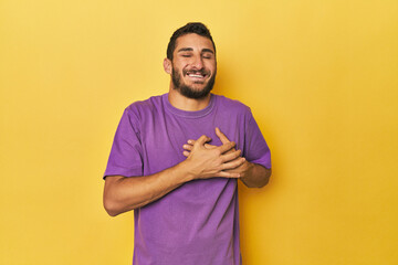 Young Hispanic man on yellow background laughing keeping hands on heart, concept of happiness.