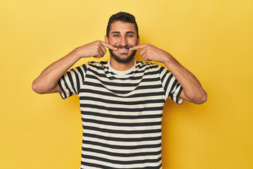 Young Hispanic man on yellow background smiles, pointing fingers at mouth.