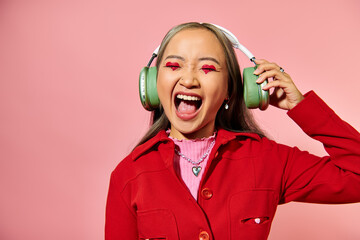 excited young asian woman listening music in wireless headphones and screaming on pink background