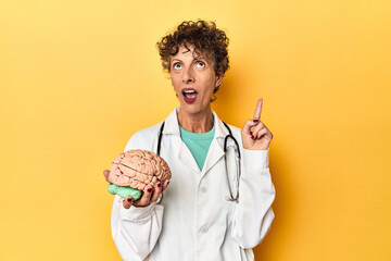 Doctor holding a brain model on yellow studio pointing upside with opened mouth.