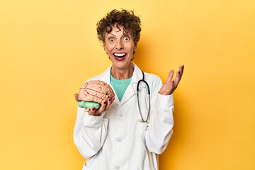Doctor holding a brain model on yellow studio receiving a pleasant surprise, excited and raising...