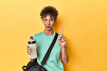 Sportswoman with gym bag & water on yellow studio showing number one with finger.