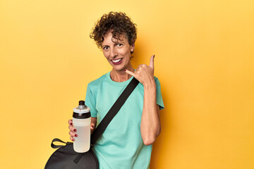 Sportswoman with gym bag & water on yellow studio showing a mobile phone call gesture with fingers.