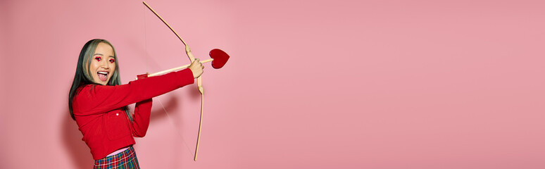 Valentines day, happy asian woman with heart shaped eye makeup holding cupid arrow and bow on pink