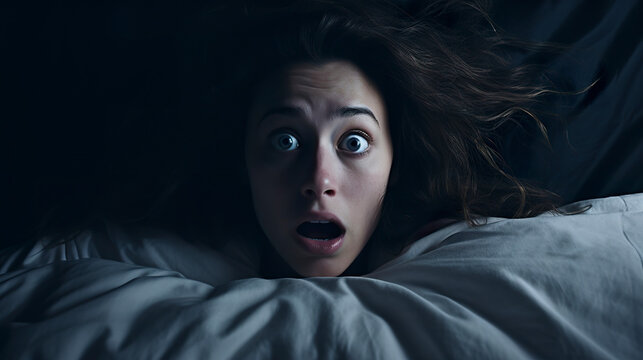A frightened young woman in bed surrounded by early morning darkness. Young woman with eyes full of fear and anxiety in the silence of the night.
