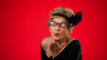 Fun smiling elderly woman 80s years old walks into frame smiling, blows a kiss and walks off frame, no teeth isolated on red background. Portrait of emotions of love