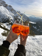 
A couple holds glasses of Aperol in glass goblets with straws on a mountain top. Panoramic views of mountains and snow. Hands of lovers on the mountain.