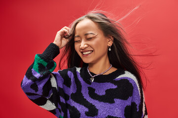 wind in hair of positive young asian girl in sweater with animal print posing on red background