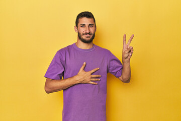 Young Hispanic man on yellow background taking an oath, putting hand on chest.