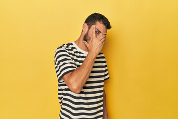 Young Hispanic man on yellow background blink at the camera through fingers, embarrassed covering face.