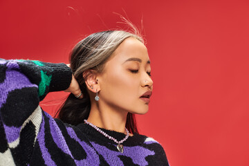 alluring young asian woman in vibrant sweater with animal print adjusting hair on red backdrop - Powered by Adobe