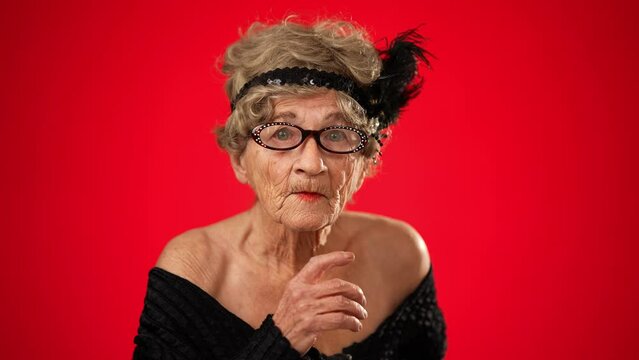 Portrait of funny looking angry upset frustrated elderly senior old woman saying shh, quiet, with wrinkled skin and grey hair on red background.