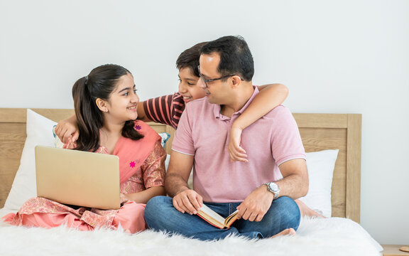 Indian single father, teenage son, daughter using laptop, sitting on bed at home in holiday, watching movie, online learning, doing homework together, happily smiling. Education, Family Concept.
