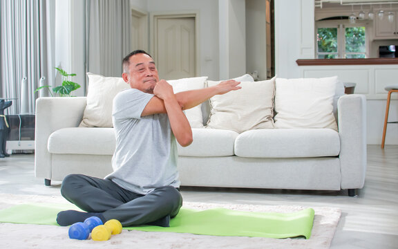 Asian aging senior old man wearing casual comfortable t shirt, sitting on floor in living room at cozy home, doing exercise with happiness, smiling. Healthcare, Retirement Concept.