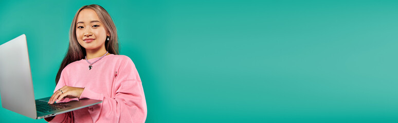 happy asian woman in pink sweatshirt using laptop while standing on turquoise backdrop, banner