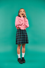 amazed young asian woman in pink sweatshirt talking on smartphone on turquoise backdrop