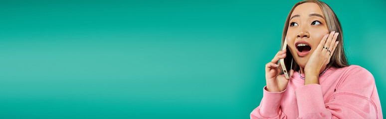 portrait of shocked young asian woman in pink sweatshirt talking on smartphone on turquoise, banner