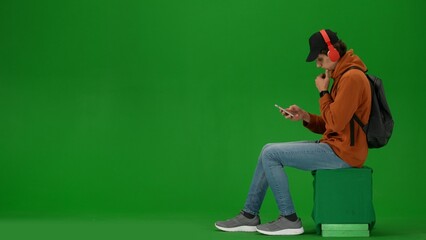 Portrait of person tourist isolated on chroma key green screen background. Young man sitting holding smartphone listening music in headphones.