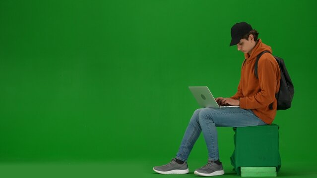 Portrait of person tourist isolated on chroma key green screen background. Young man sitting holding laptop and working, waiting for flight.