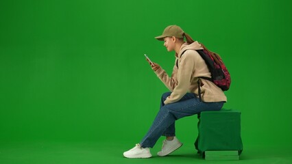 Portrait of person tourist isolated on chroma key green screen background. Young woman sitting holding smartphone looking at screen smiling expression.