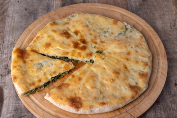 Pie with spinach, herbs and suluguni cheese on a wooden board. Whole pie with cut off piece. Traditional round flat pie with filling. Homemade pastries of Caucasian and Balkarian cuisine. 
