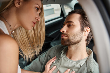 passionate blonde woman seducing bearded man sitting in car on urban street, urban romance