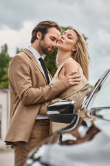 businessman in formal wear embracing passionate blonde woman near car on urban street, passion