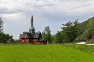 Fototapeta na wymiar Green field and the historical Kvikne kirke, with its towering spire, surrounded by lush trees in Kvikne. The scene is enhanced by a beautifully curved roadway to the right. Innlandet, Norway