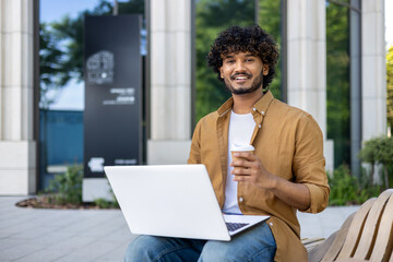 Portrait of a young Indian freelancer man sitting on a bench outside with a laptop, holding a cup...