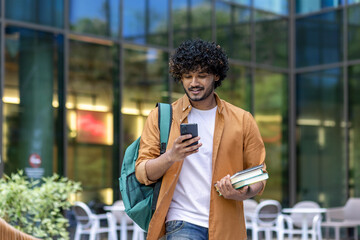 Indian young male student walking on street near campus and university with backpack. Holds books and uses a mobile phone