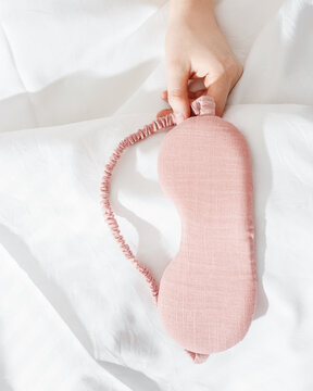 Pink eye mask for sleep in woman hand on white bedclothes, minimal lifestyle aesthetic photo. Top view Female sleeping mask for best sleepers, for travel, comfort relax. Rest well concept.