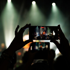 Person recording a concert with their phone. The stage is visible on the phone screen and is lit up. - obrazy, fototapety, plakaty