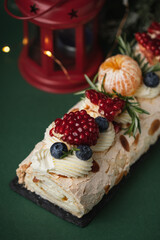 Meringue roll sprinkled with powdered sugar decorated with blueberries, pomegranate and almond flakes on the dark green background with blurred Christmas tree. Christmas festive pastry