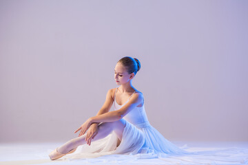 elegant ballerina in pointe shoes sits with her legs stretched out in a long white skirt on a white...