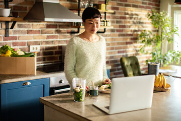 Woman preparing smoothie with laptop in home kitchen