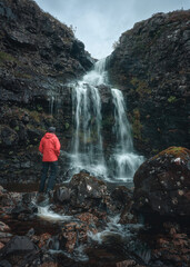 A hiker in a red jacket looks at a waterfall on the bank of a mountain stream. View of a waterfall on a mountain stream. Isle of Skye, Scottish Highlands, UK