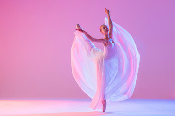 a young ballerina in pointe shoes dances in a long flowing white skirt on a pink background