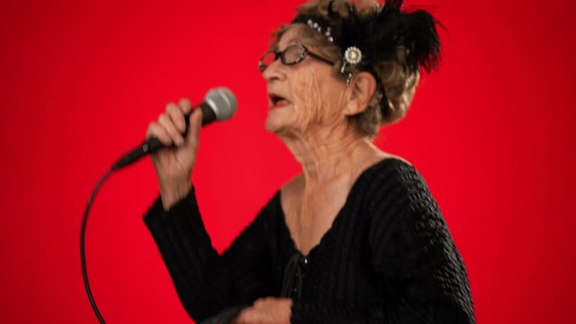 Funny toothless elderly woman in fashionable look with glasses, walks into frame singing enthusiastically into a microphone and dancing isolated on solid red background
