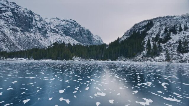 Aerial view from low flying drone of frozen lake in snowy mountains in winter at sunset. Lofoten islands, Norway. Top view of frosted river, reflection on ice, snowy rocks, sky. Cloudy weather. Nature
