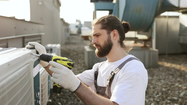 Bearded craftsman crouching and using drill while repairing air conditioner on roof of factory. Caucasian skilled male dressed in uniform servicing and improving machine equipment.