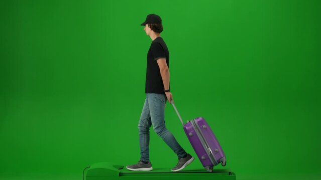 Portrait of traveler isolated on chroma key green screen background. Young man with suitcase in cap walking and looking around.
