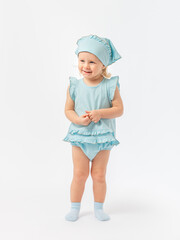 A blonde girl of 2 years old is smiling, touching her fingers and looking away in full height in a blue dress and socks on a white background.