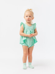 A thoughtful, serious 2-year-old toddler girl stands with her hands clasped on a white background in a green bodysuit dress.