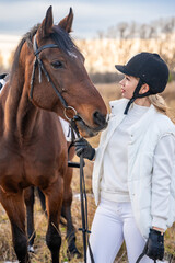Portrait of blond professional female jockey standing near horse. Friendship with horse