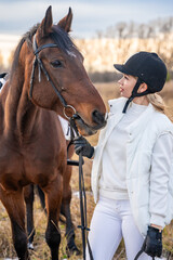 Fototapeta premium Portrait of blond professional female jockey standing near horse. Friendship with horse